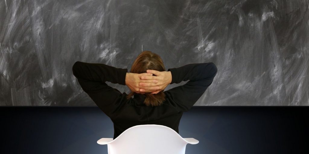 Persona sentada de espaldas, con las manos detrás de la cabeza frente a una pizarra.