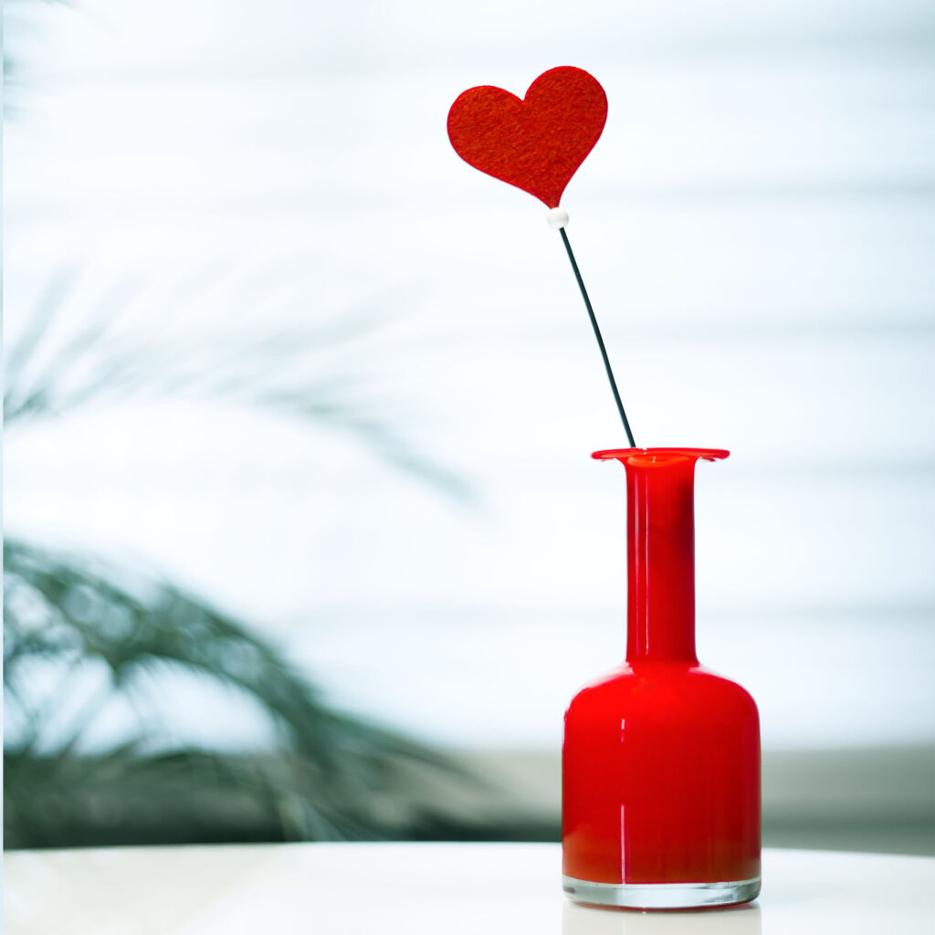 Un corazón rojo decorativo sobresale de un jarrón rojo sobre un fondo claro.