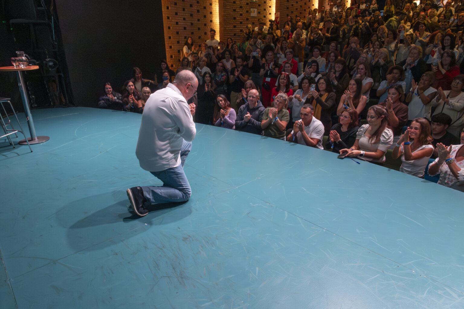 Un ponente se arrodilla en el escenario mientras el público lo aplaude.